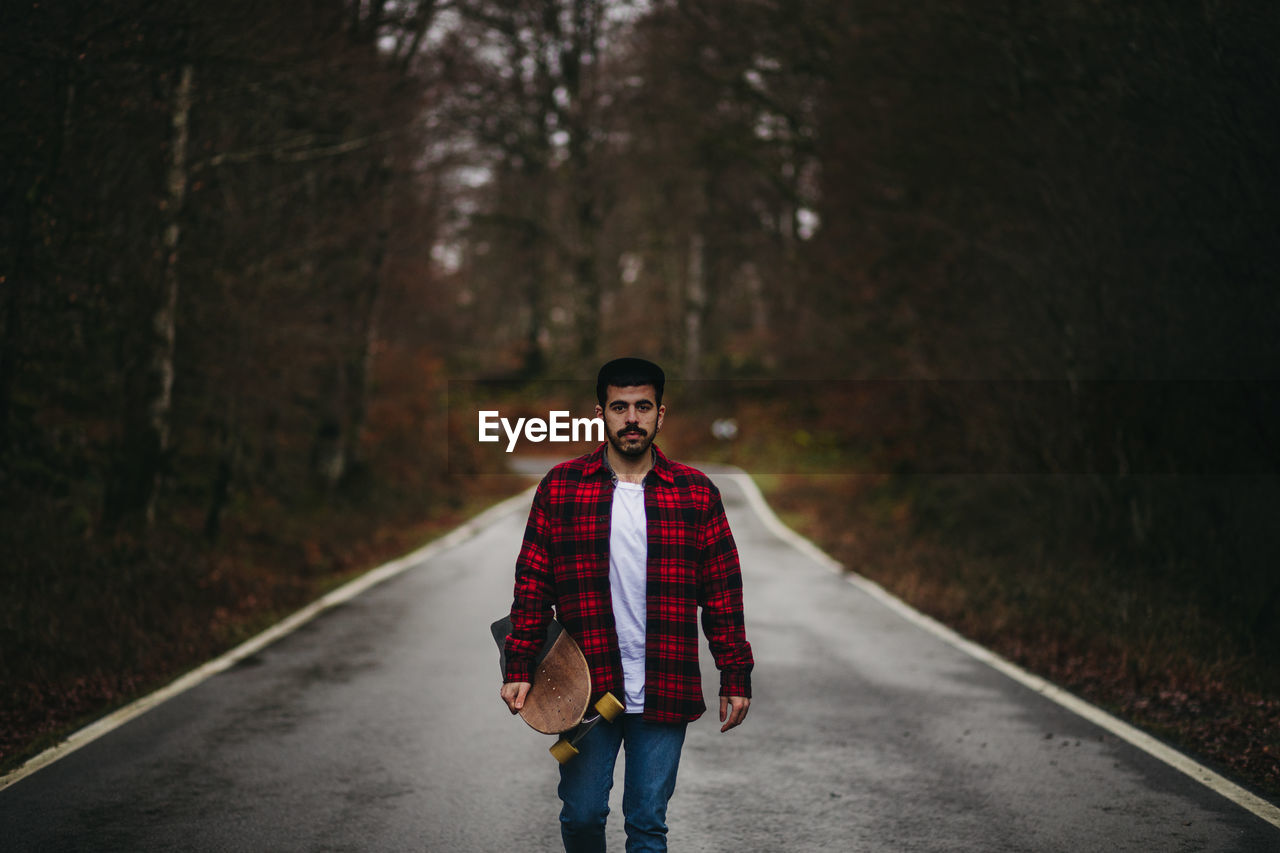 Young stylish man in casual wear walking on asphalt road with skateboard in hand on autumn day looking at camera