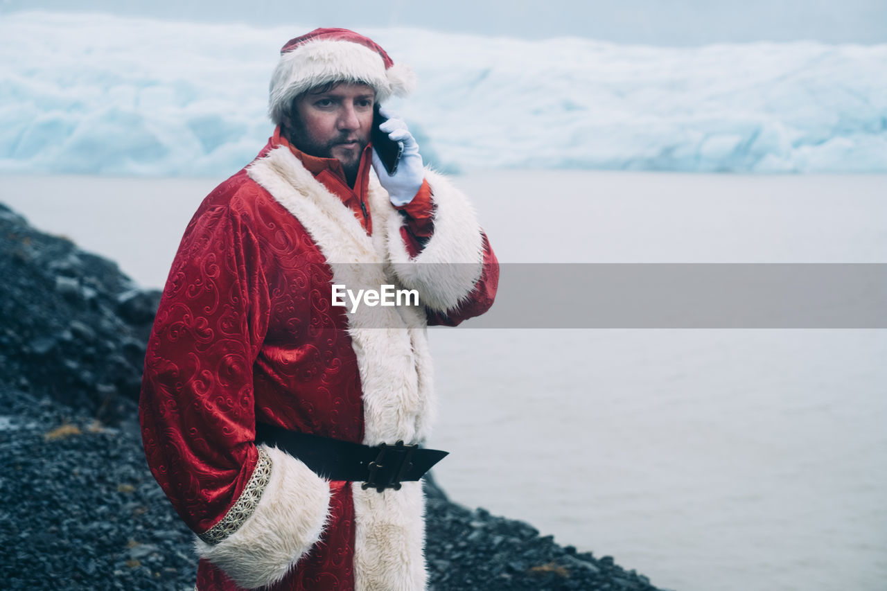 Man in santa costume talking on smart phone while standing by lagoon during winter