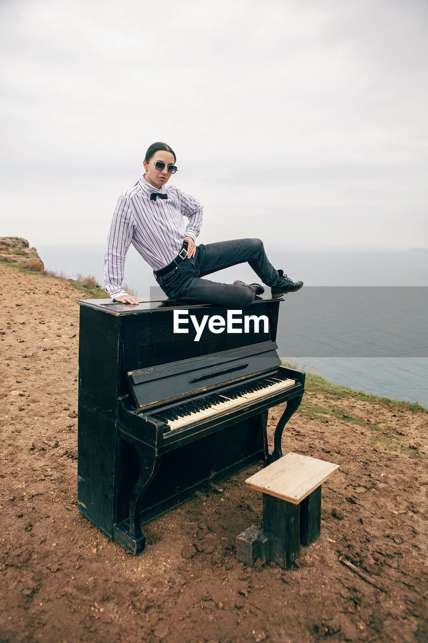 Woman in sunglasses and a white shirt sits on a black piano that stands on cape fiolent 