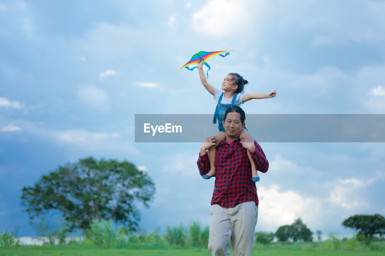 Father giving piggy back ride to daughter holding kite against cloudy sky