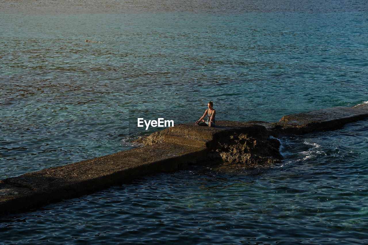 Peaceful female sitting on stone in padmasana and meditating with mudra gesture near sea during sundown