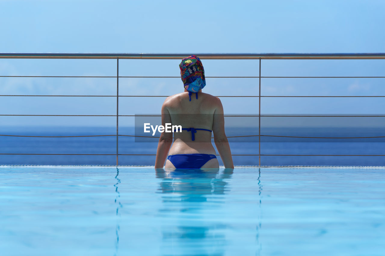 Rear view of bikini woman looking at sea while standing in infinity pool