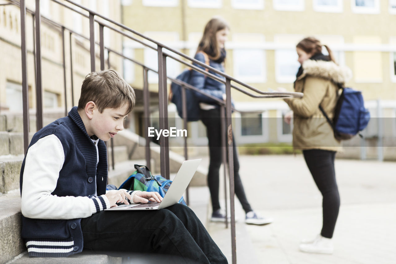 Boy using laptop on steps with schoolgirls in background outside school building