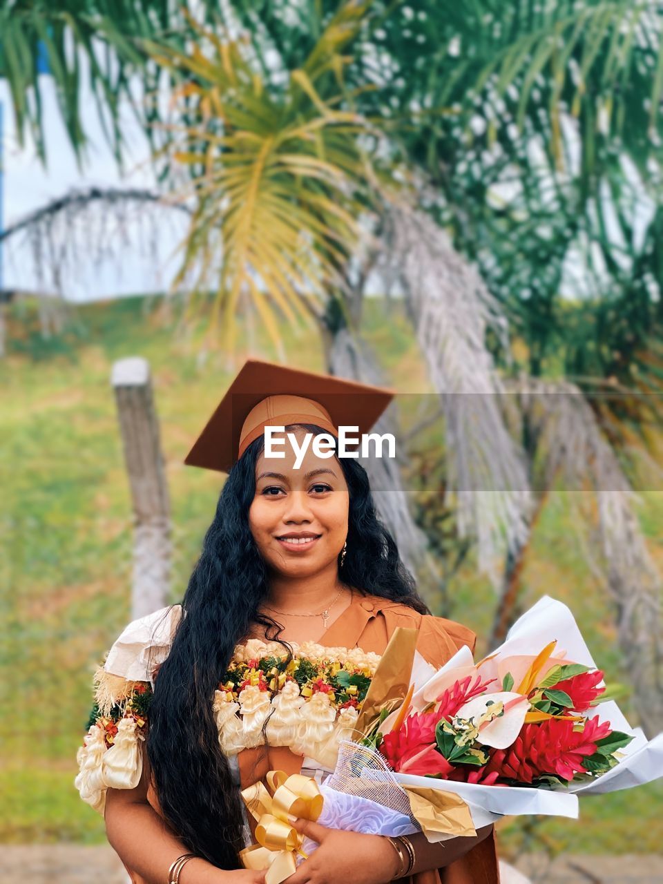 Portrait of smiling young woman in graduation gown standing against trees