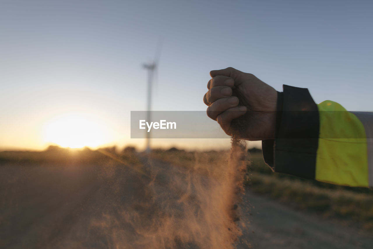 Close-up of man's hand scattering soil at a wind turbine at sunset