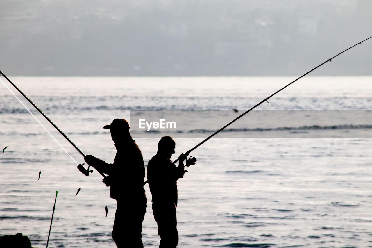 SILHOUETTE MAN FISHING AT SEA SHORE AGAINST SKY