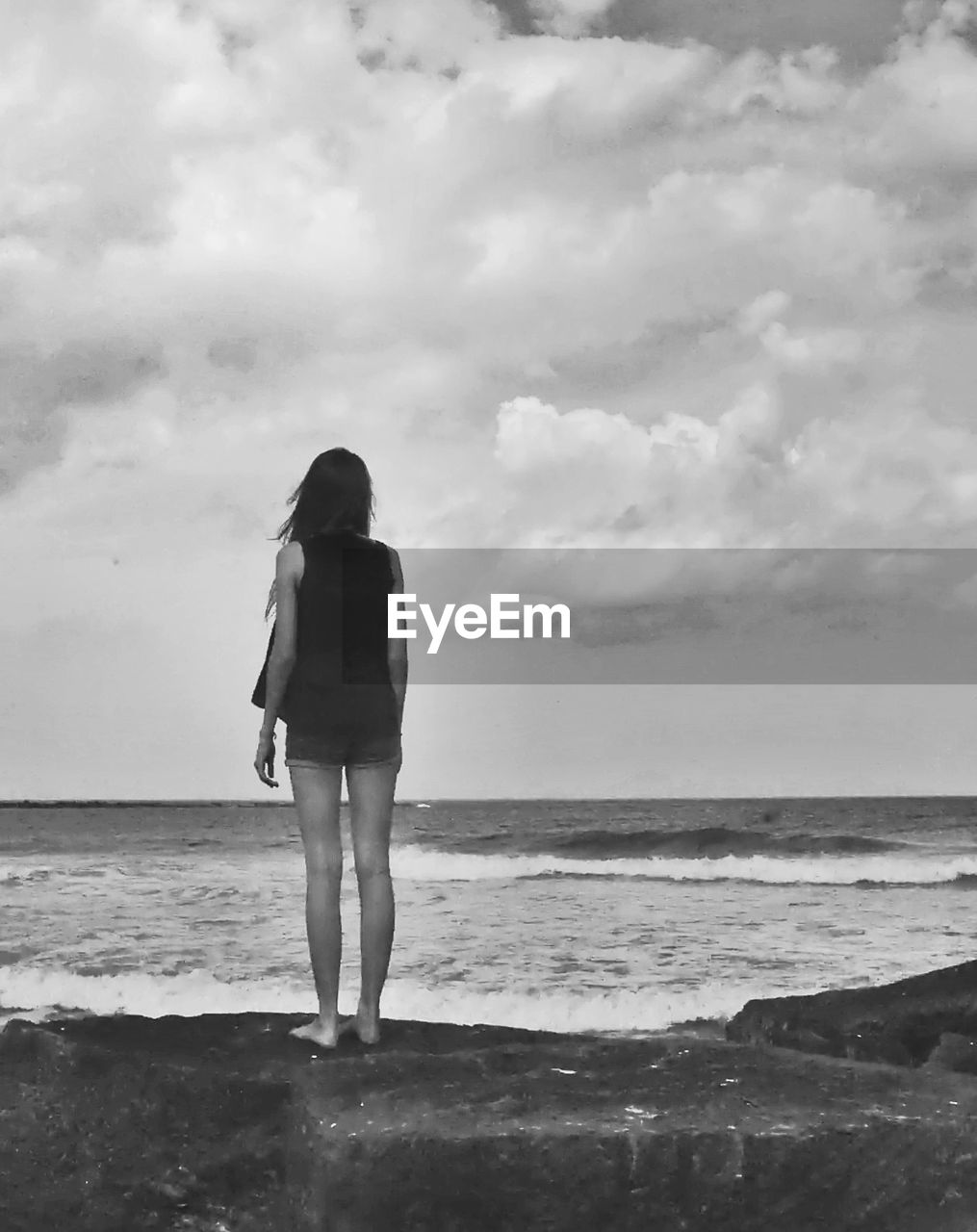 Rear view of young woman standing on rock in front of sea against cloudy sky