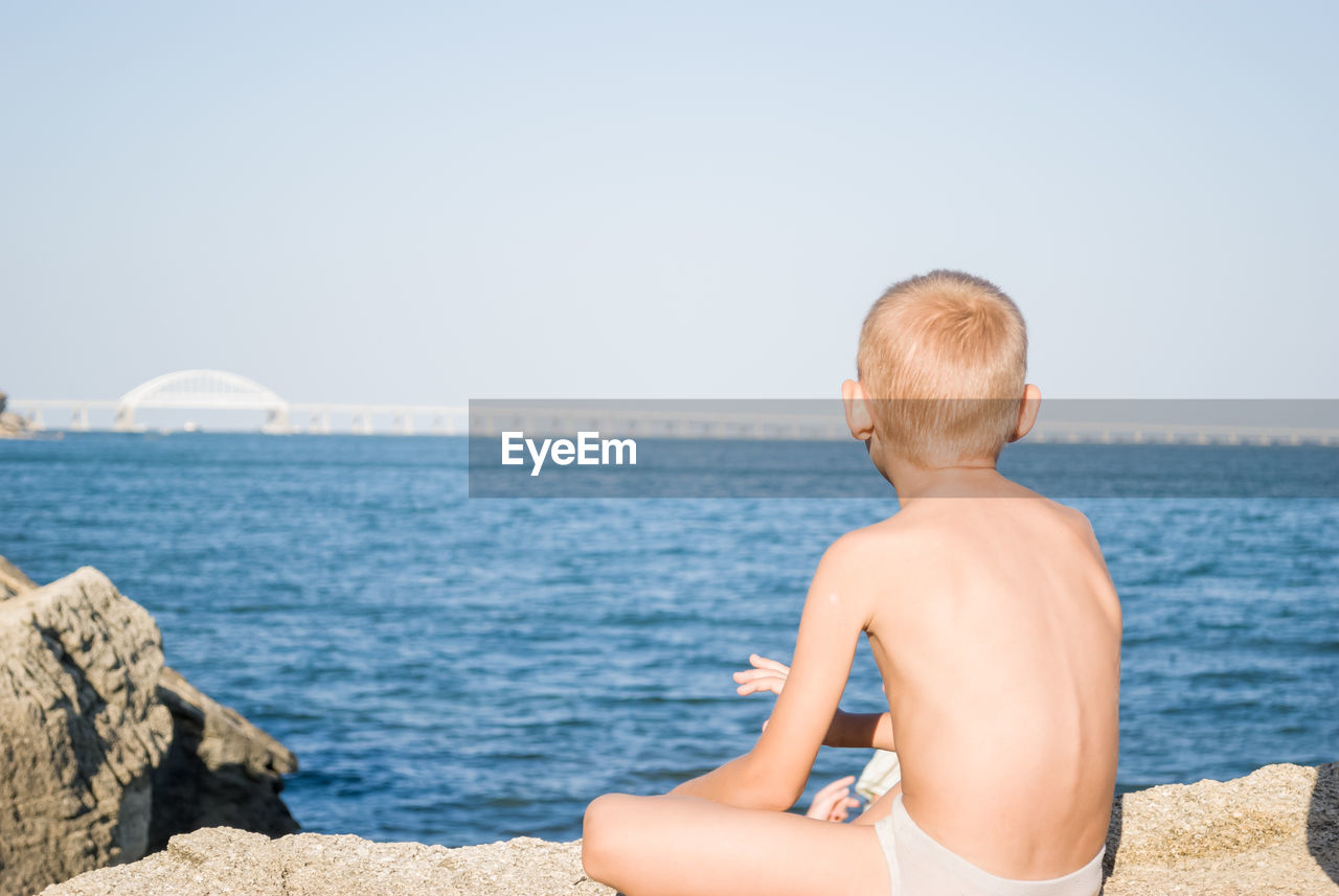 Rear view of boy sitting on rock by sea against sky