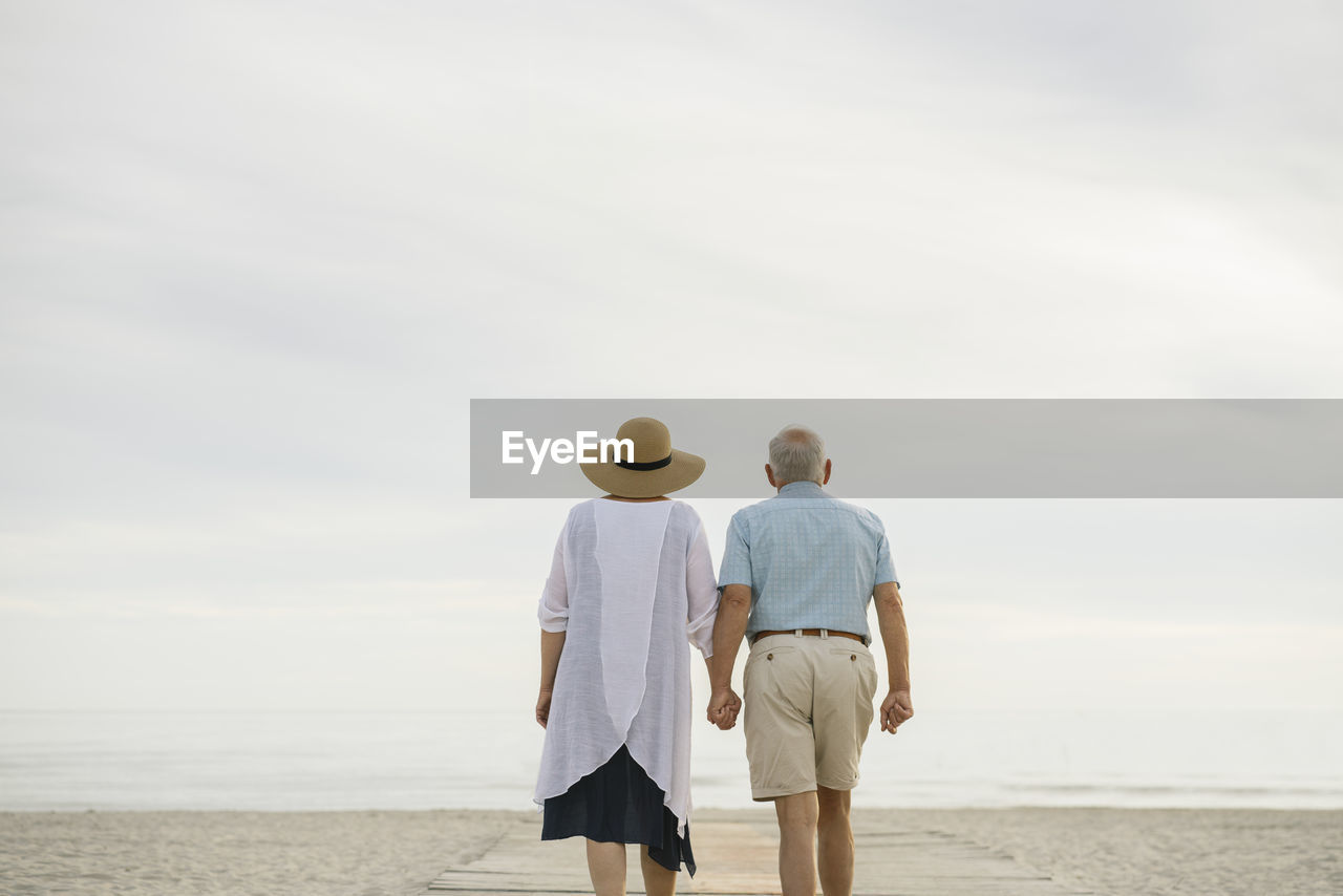 Back view of senior couple standing hand in hand on wooden boardwalk looking to the sea, liepaja, latvia