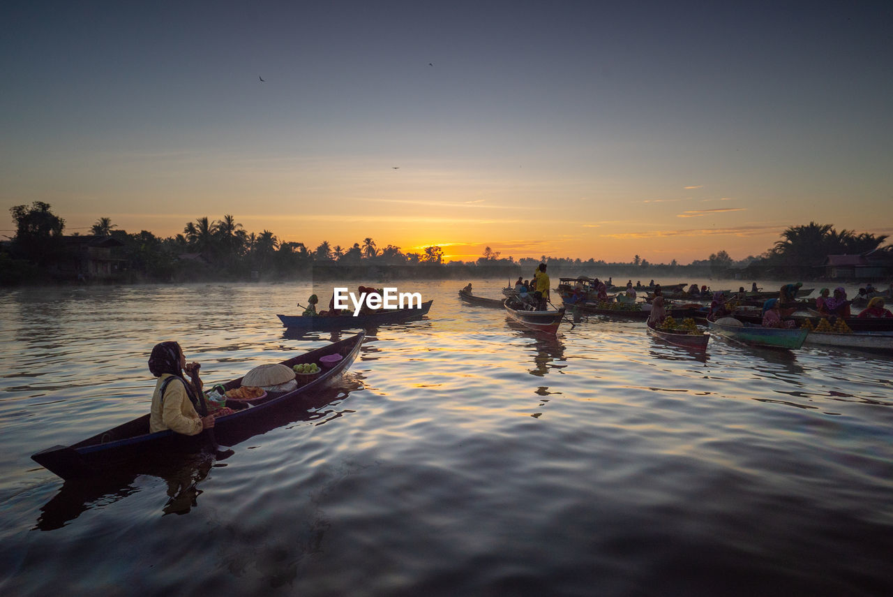 People in boats on river against sky during sunset