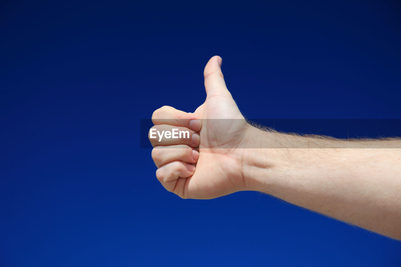 Cropped hand of man showing thumbs up against clear blue sky