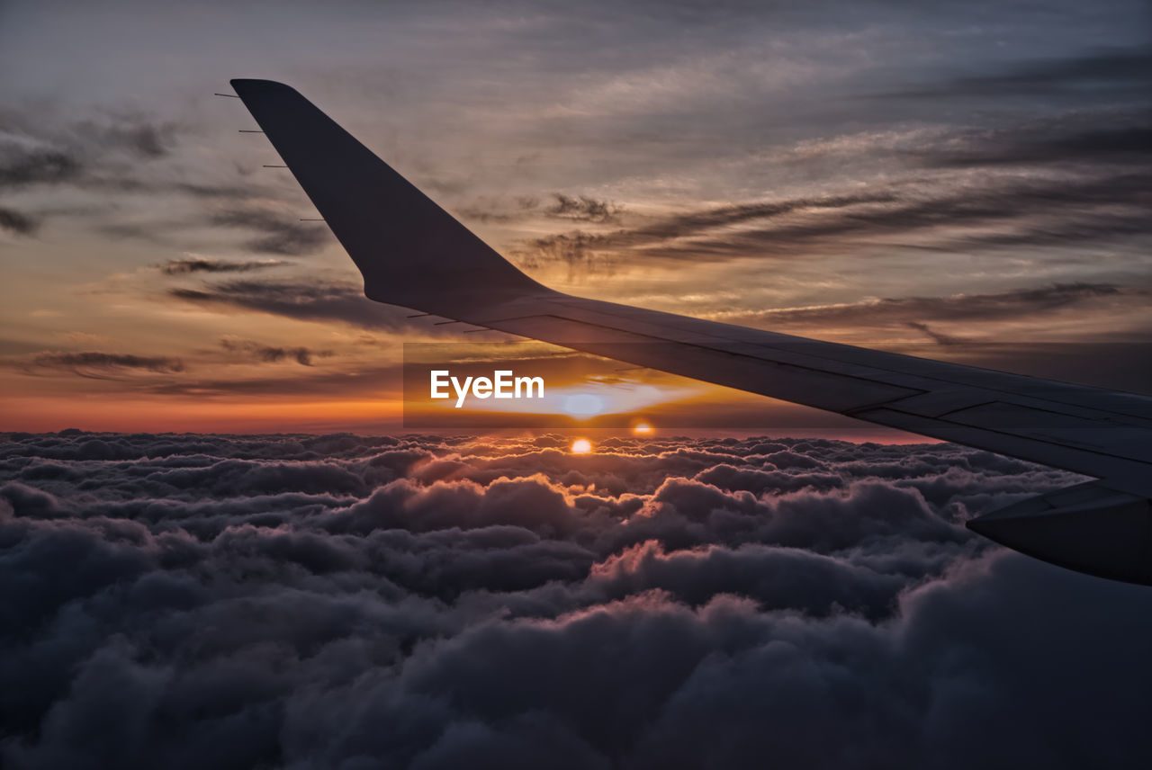 Cropped image of aircraft wing flying over cloudscape against sky during sunset