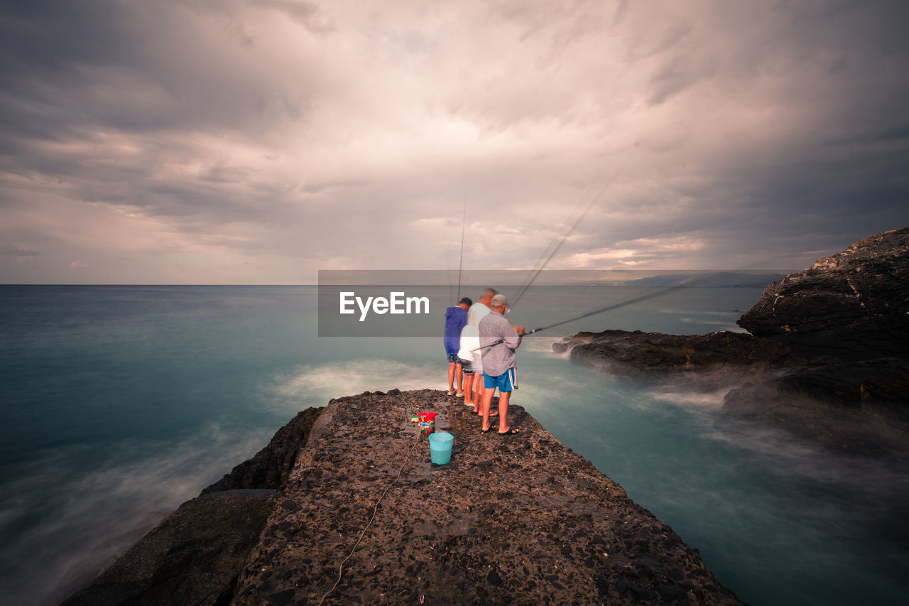 Men fishing while standing on rock in sea against cloudy sky