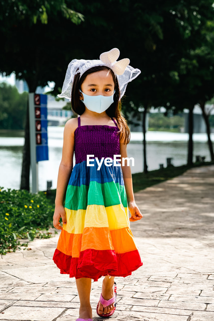 Little girl wearing face mask with rainbow dress enjoying outdoor in the park. new normal.