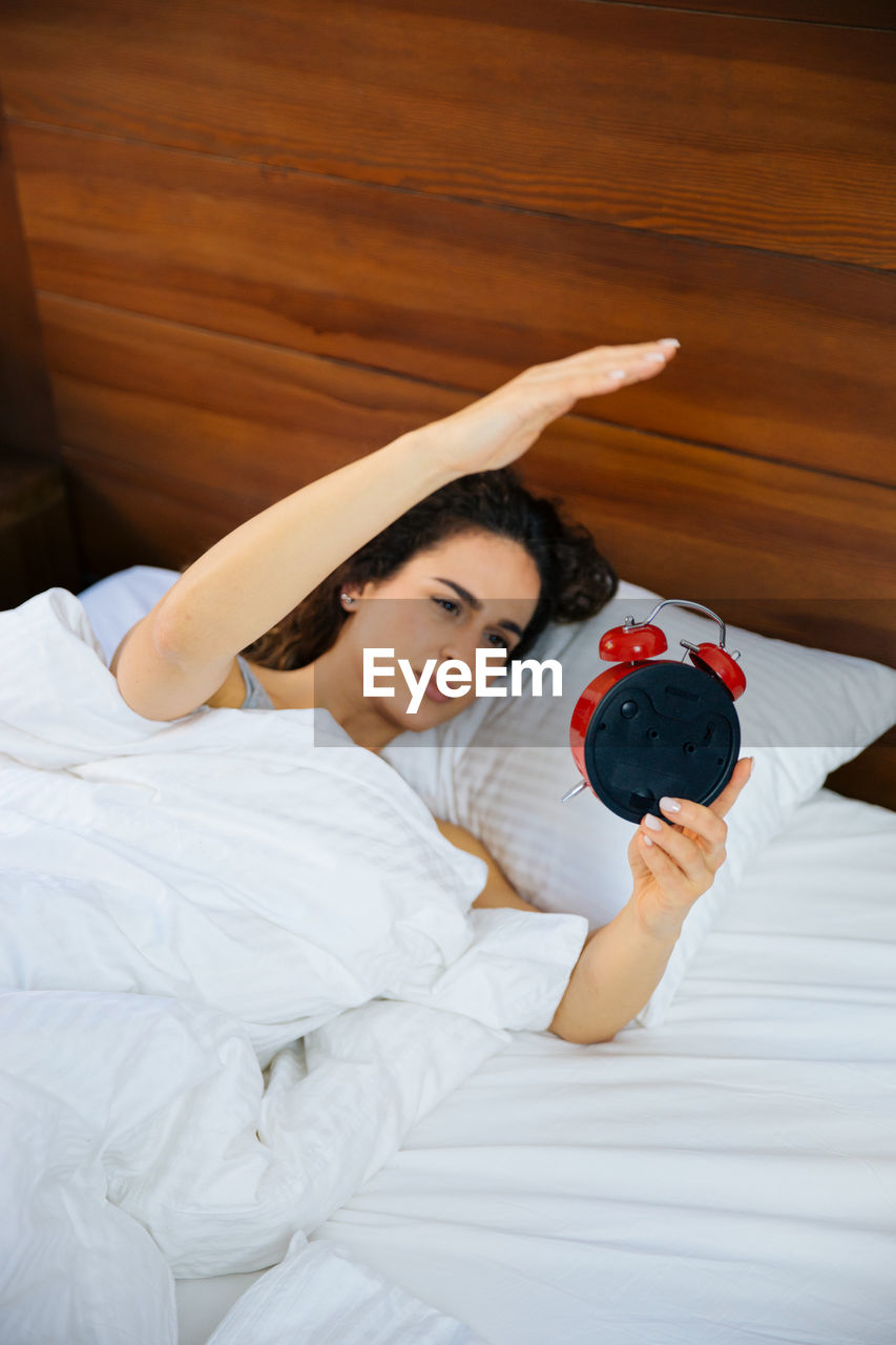 High angle view of young woman holding alarm clock while sleeping on bed at home
