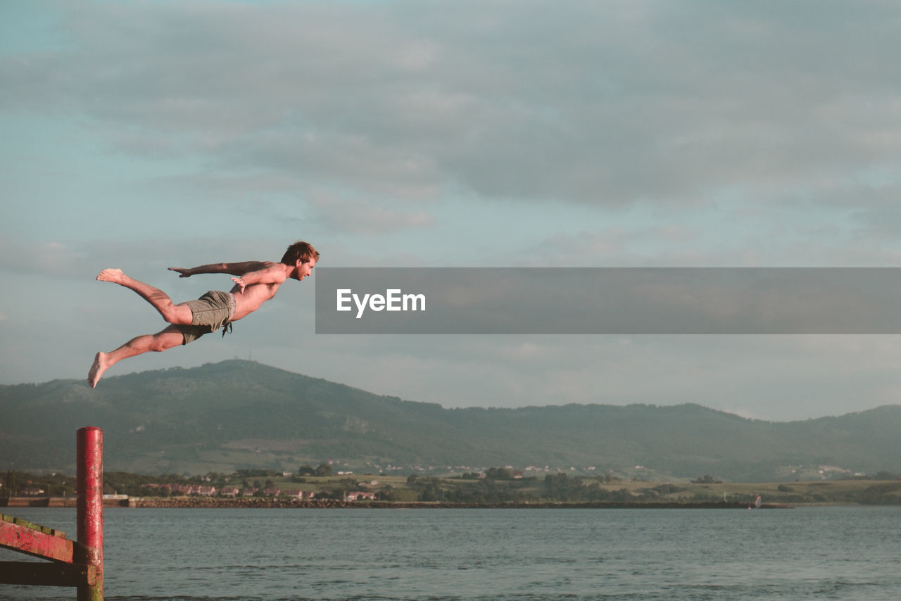 WOMAN JUMPING OVER SEA AGAINST MOUNTAINS