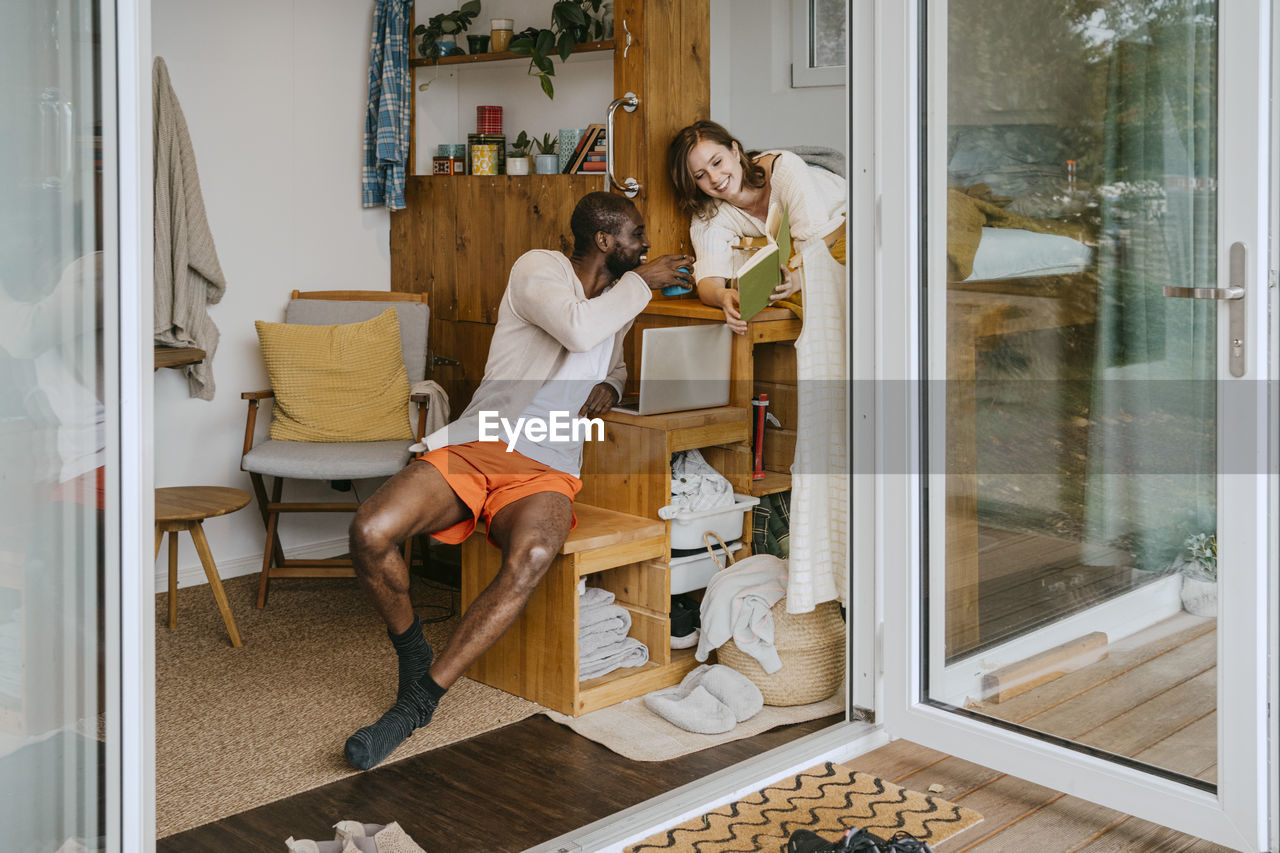 Smiling woman sharing book with man sitting on seat at home seen through doorway