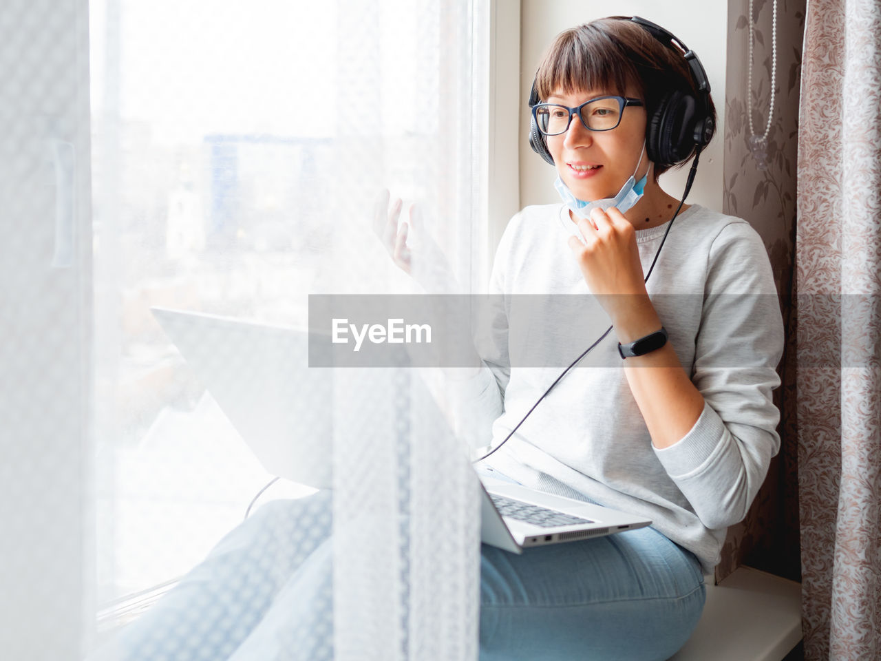 Woman in medical mask works remotely from home. she speaks online with somebody with laptop.