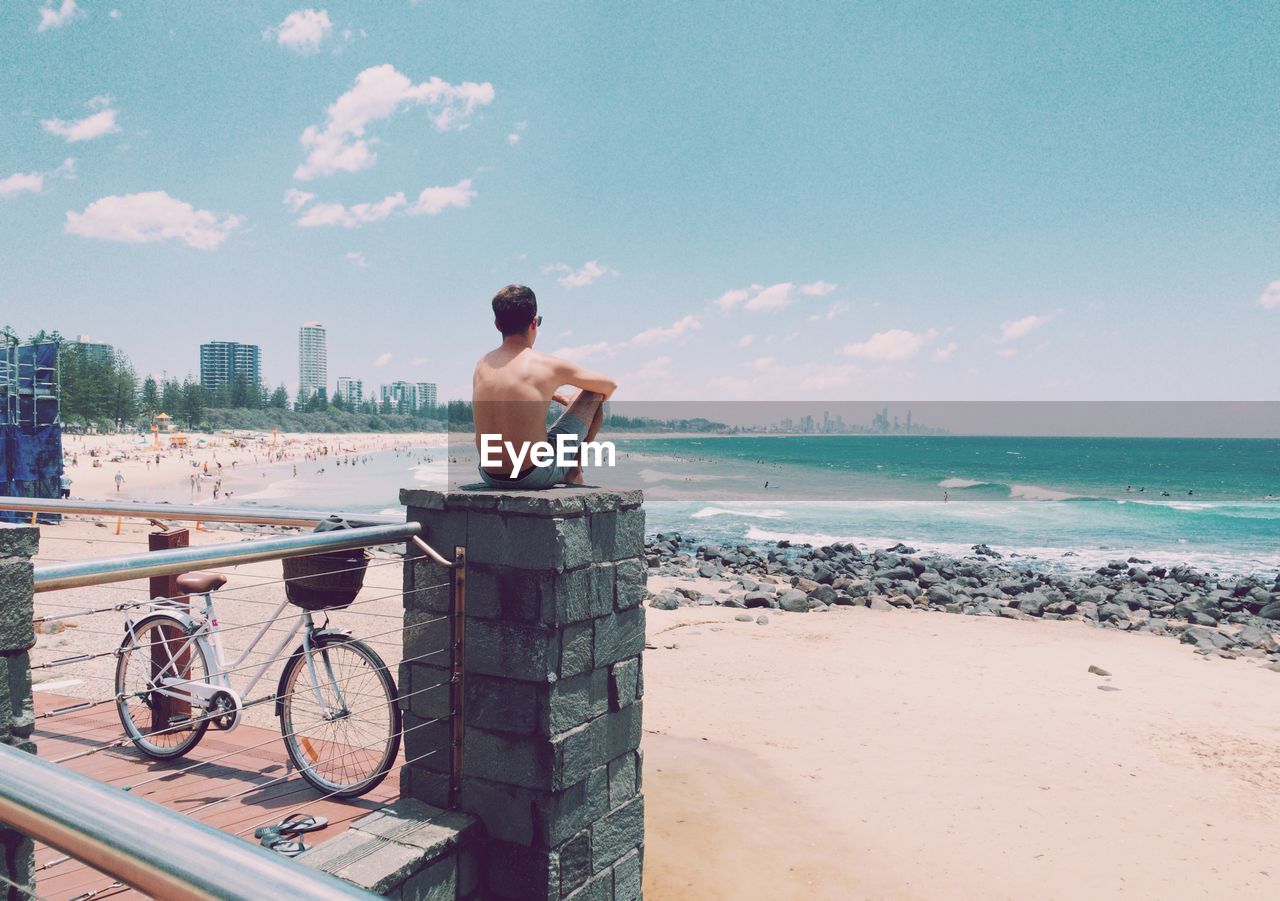 REAR VIEW OF SHIRTLESS MAN STANDING ON BEACH