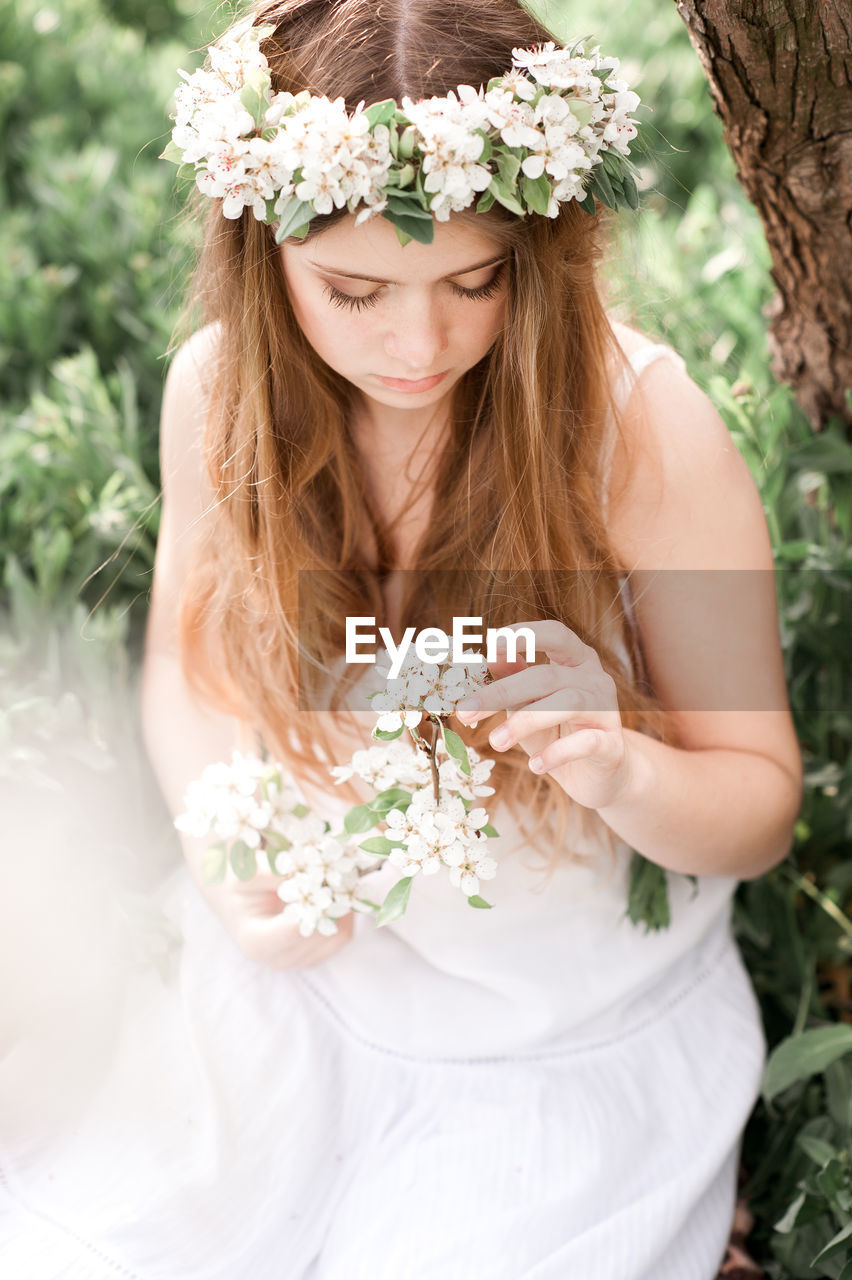 bride, women, plant, wedding dress, flower, one person, long hair, flowering plant, clothing, adult, young adult, hairstyle, dress, nature, female, wreath, laurel wreath, happiness, wedding, celebration, event, emotion, newlywed, portrait, smiling, fashion, front view, flower arrangement, brown hair, looking, white, bouquet, veil, fashion accessory, beauty in nature, headpiece, outdoors, crown, person, holding, tree, royalty, day, waist up, looking down, blond hair, wearing flowers, gown, standing, positive emotion, life events, summer, tiara, child, elegance, three quarter length, pink, portrait photography, freshness, redhead, headband, wavy hair