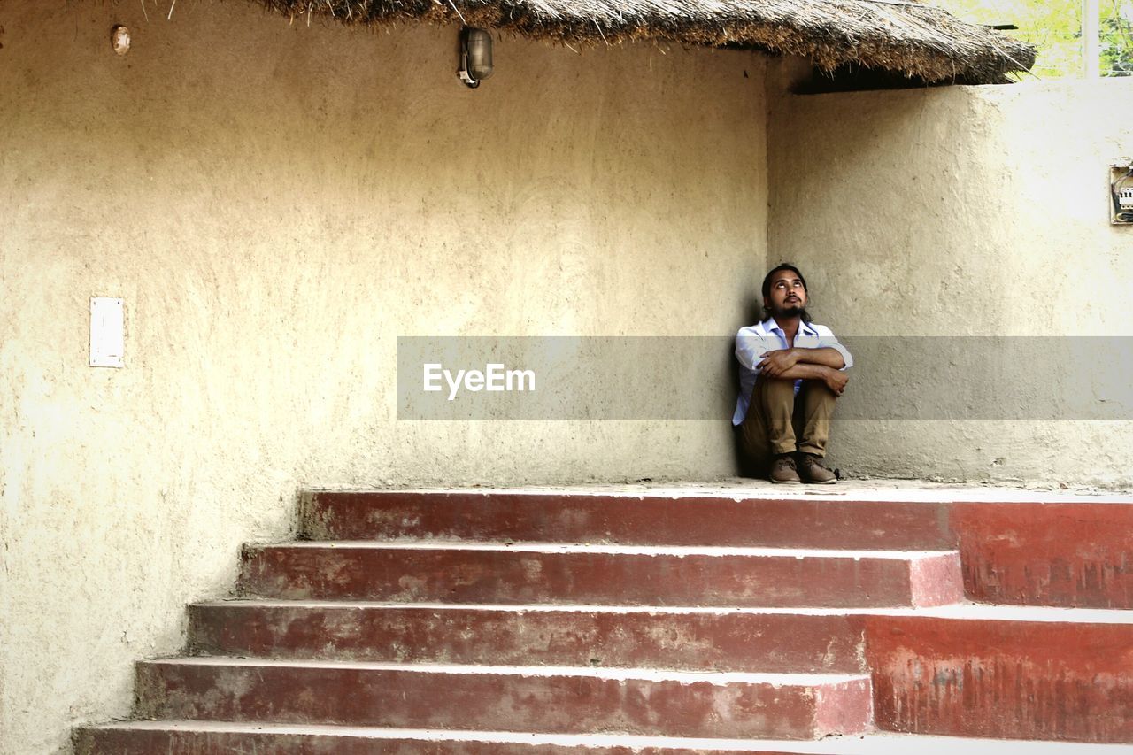 Young man looking up while sitting by house in village