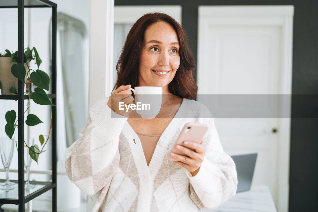 Smiling brunette woman with long hair in white cardigan using mobile phone in office