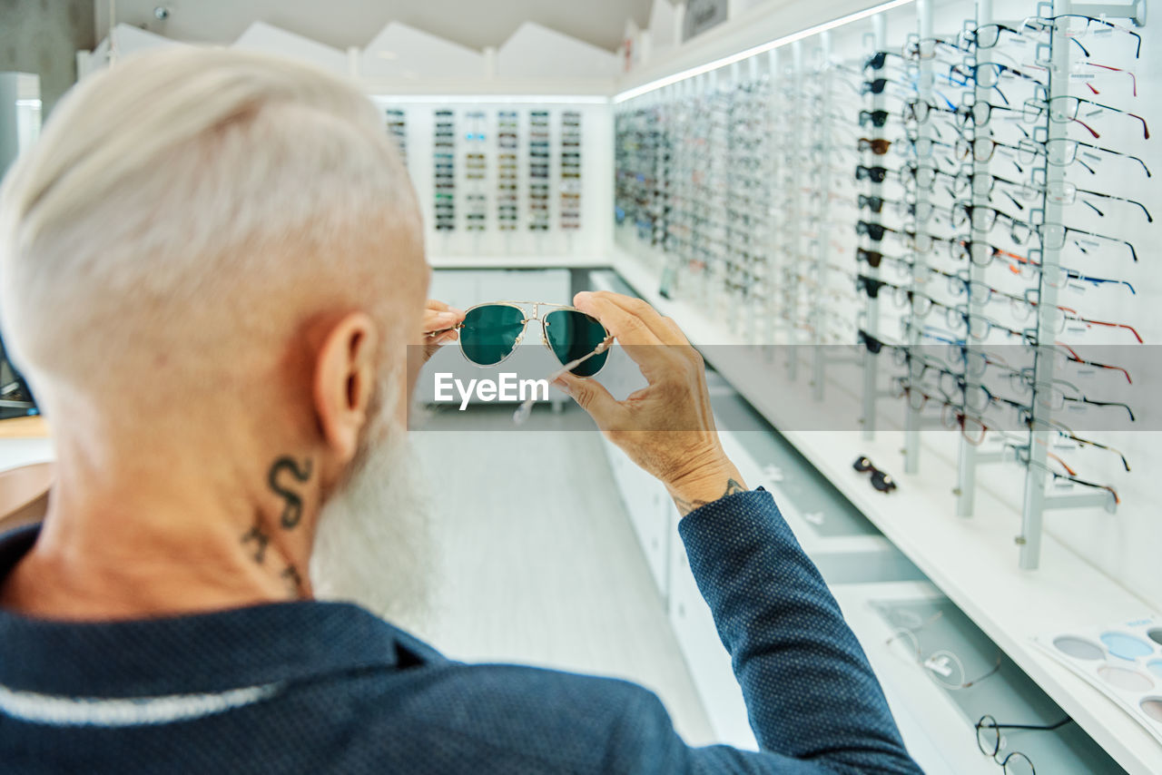 Back view elderly bearded male in trendy outfit standing with modern stylish sunglasses in optical shop