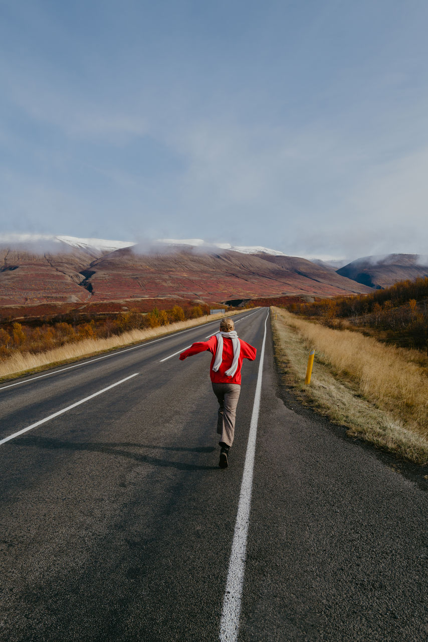 Woman in a red jumper and a scarf running on the road in iceland with arms spread