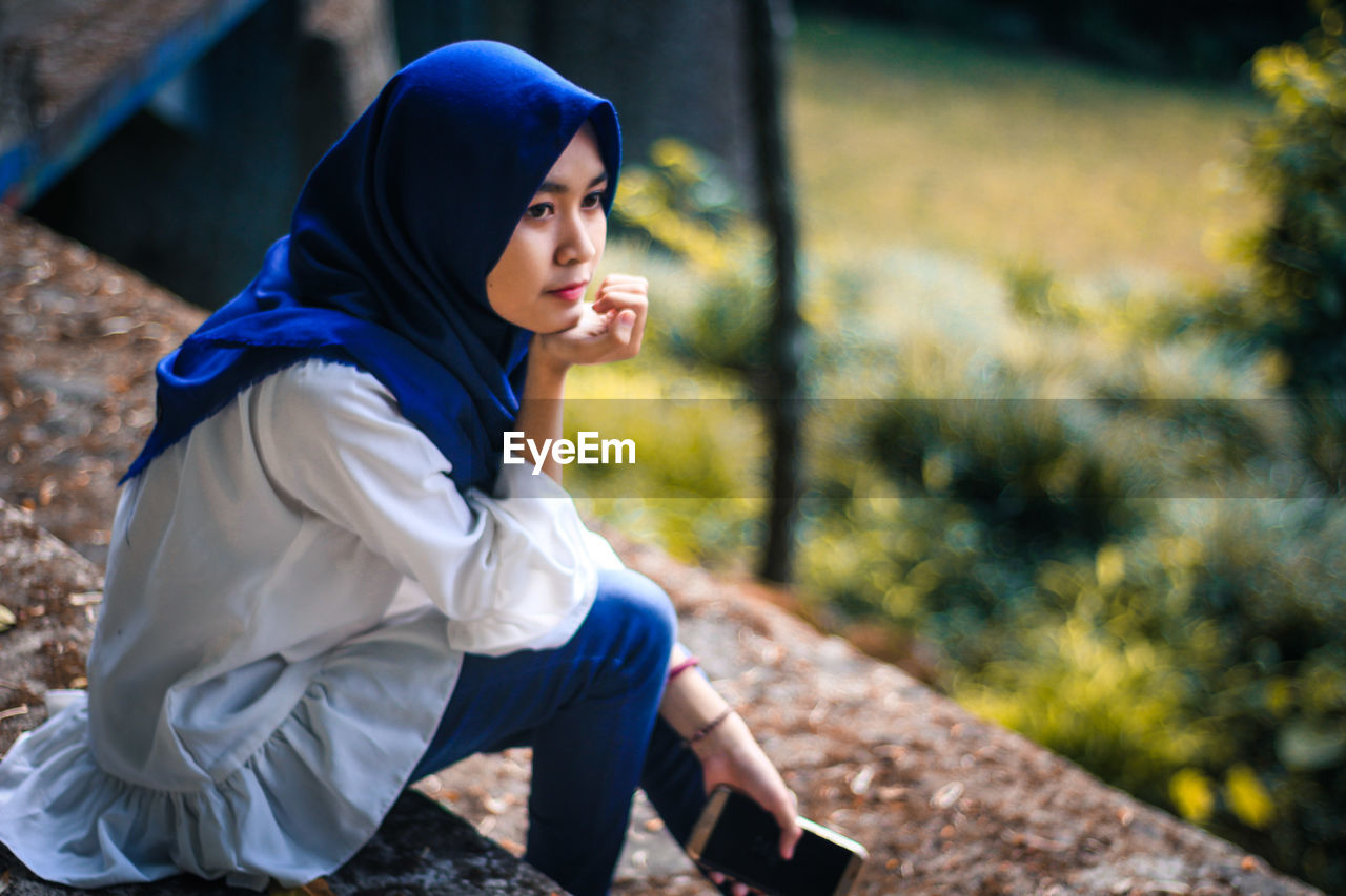 YOUNG WOMAN LOOKING AWAY WHILE SITTING ON PLANT