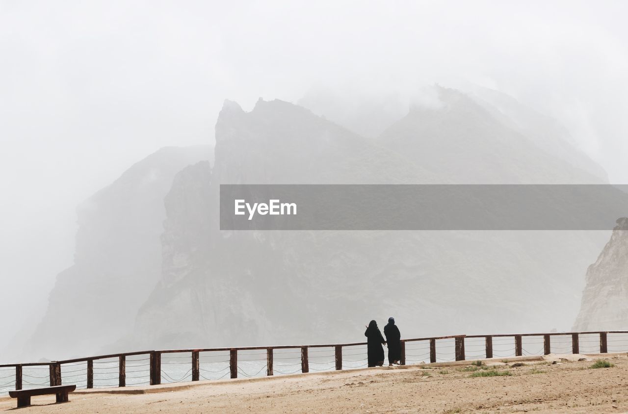 Two arabic women walking on the shore. foggy mountains in background. mughsayl / mughsail in oman.