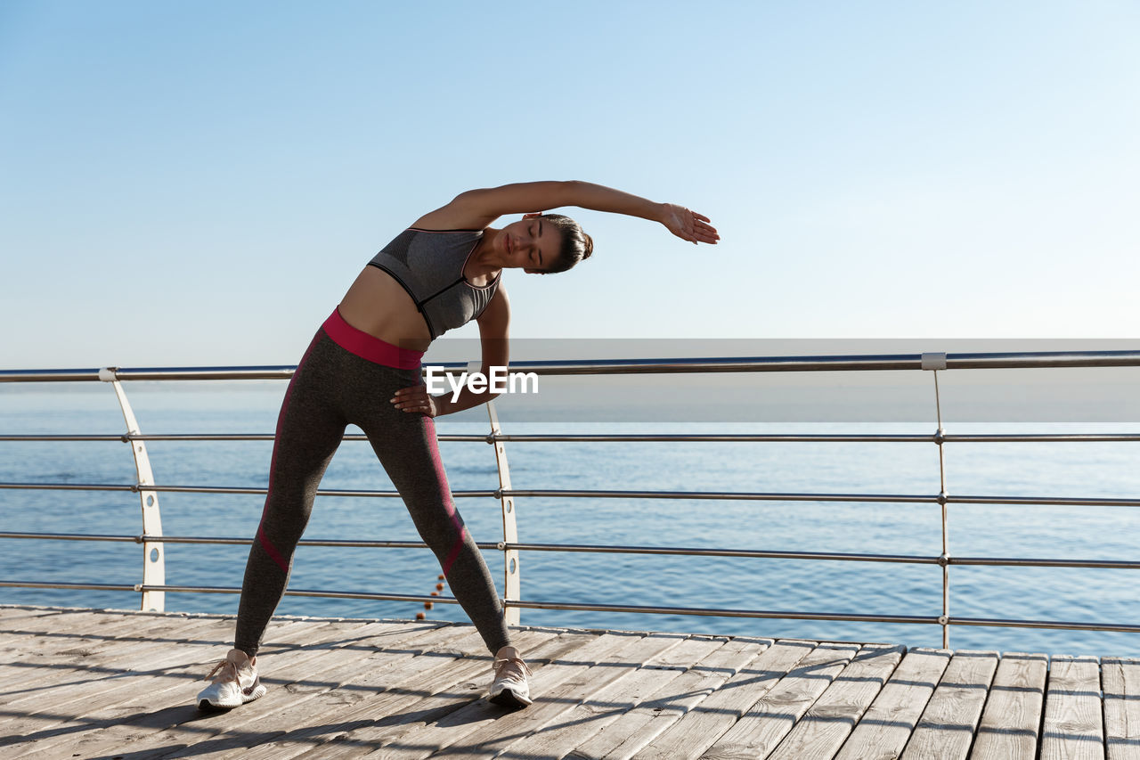 Full length of woman exercising by railing while standing against sea