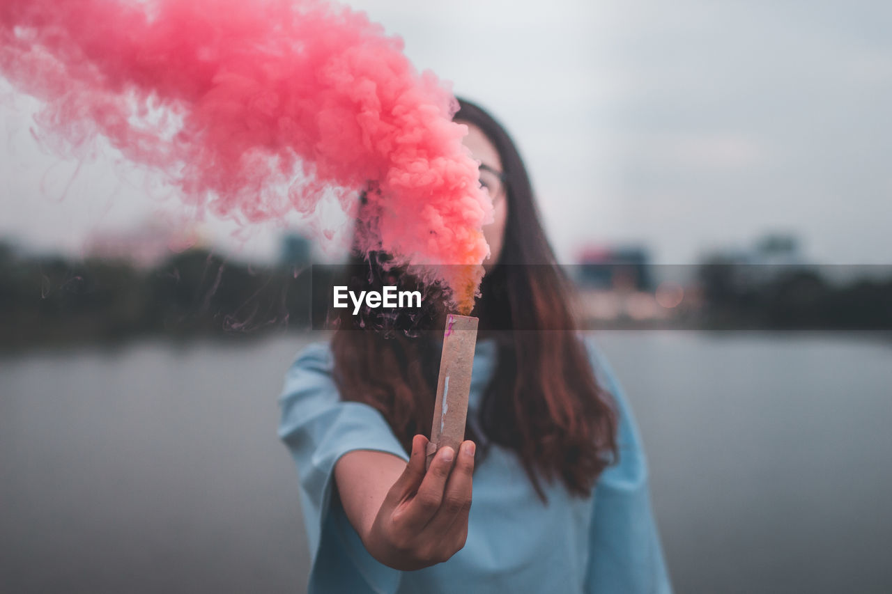 Young woman holding distress flare while standing by lake against sky