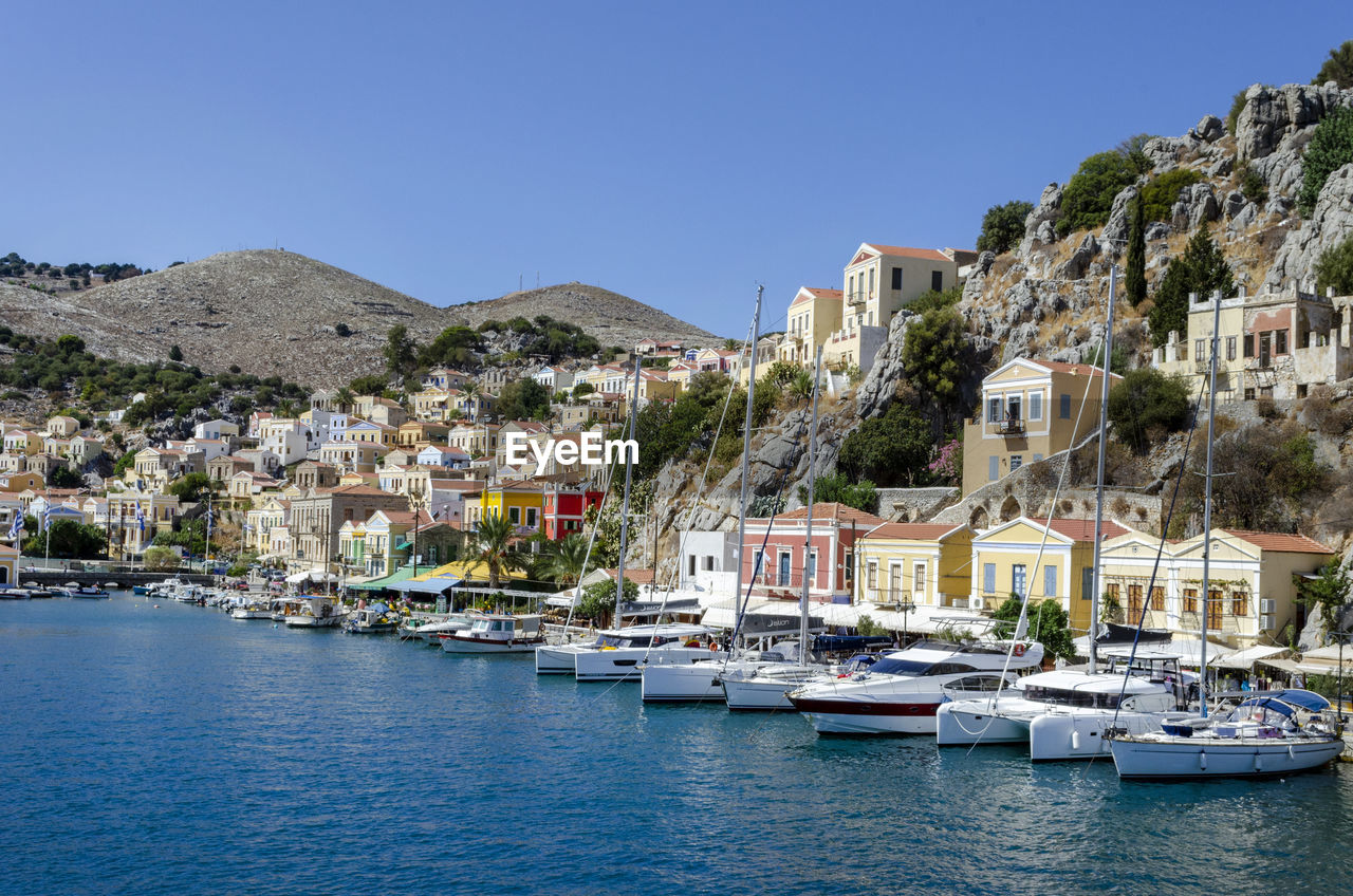 SAILBOATS MOORED ON RIVER AMIDST BUILDINGS AGAINST CLEAR BLUE SKY