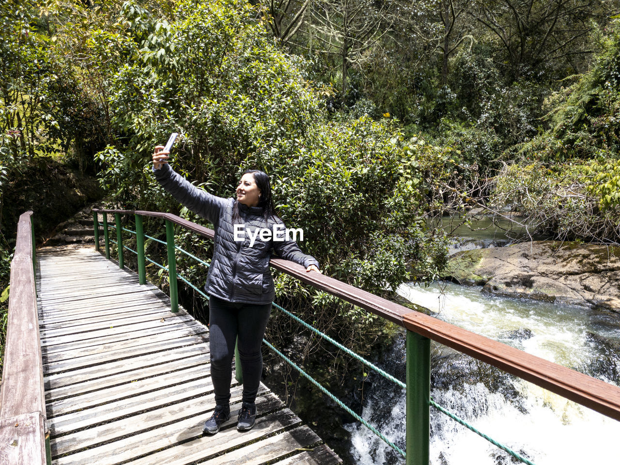 one person, full length, railing, plant, nature, tree, leisure activity, adult, standing, lifestyles, bridge, day, casual clothing, footbridge, young adult, arm, women, limb, water, outdoors, architecture, beauty in nature, wood, forest, growth, footpath, front view, sunlight, person, built structure, green, arms raised, rope bridge, female, the way forward, clothing, walkway, happiness, river, emotion, tranquility, travel, arms outstretched