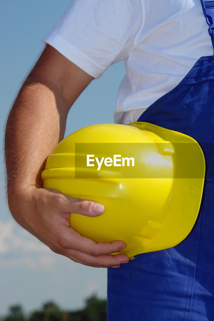 Midsection of man holding hardhat while standing against sky