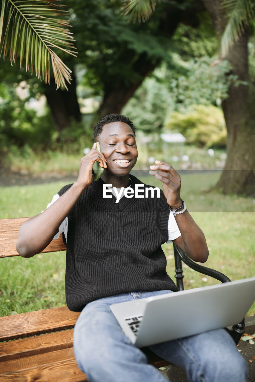 Young african man talking on phone, working on laptop in park, happy, pleased