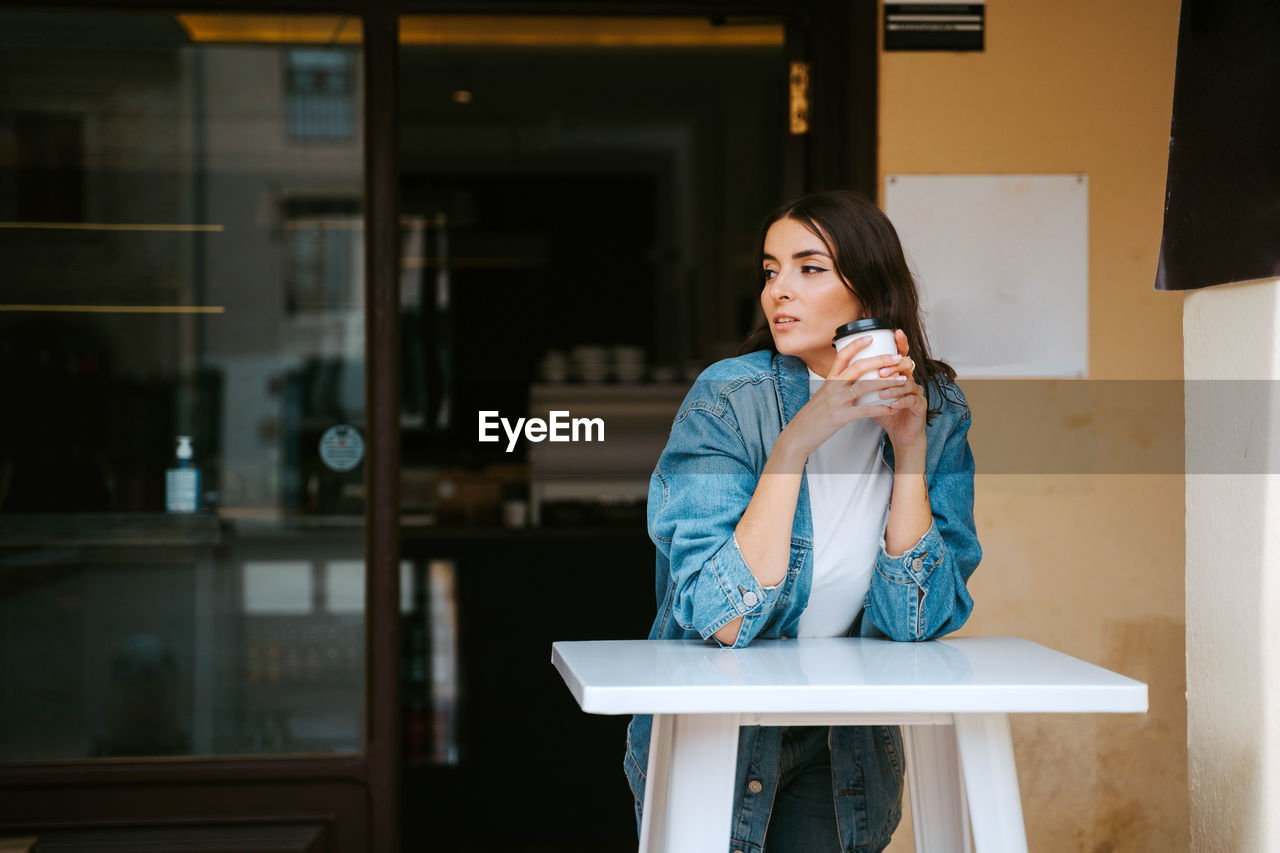 Relaxed young female in denim outfit drinking takeaway coffee while resting at table on cafe terrace