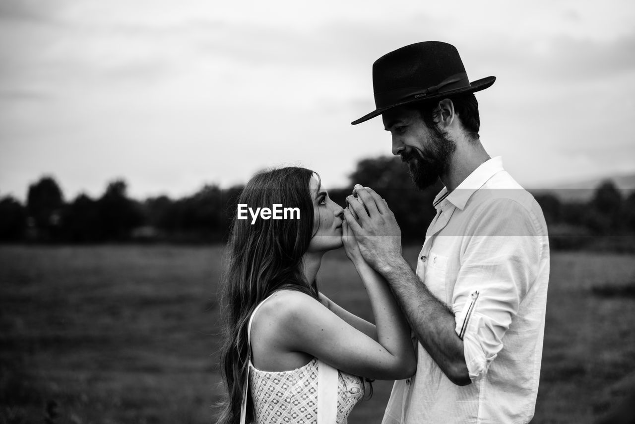 Side view of romantic couple holding hands while standing on field against sky