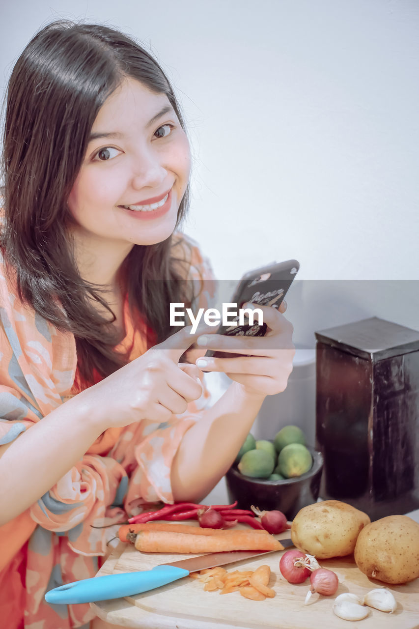 Portrait of smiling woman using phone while cutting vegetables at home