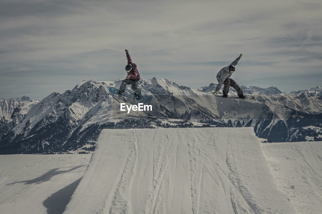 Snowboarders jumping over snow covered ramp at mountains against sky