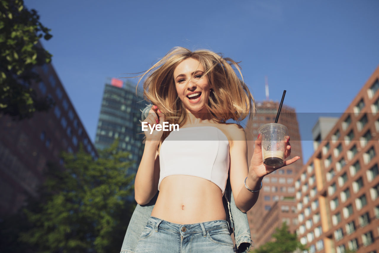 Portrait of happy woman with drink standing against buildings in city