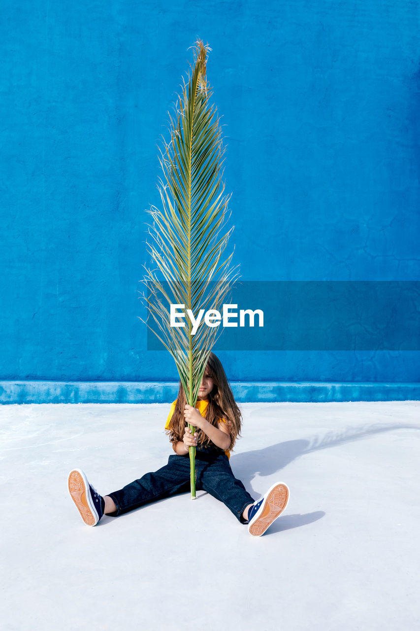 Teenager sitting with huge palm tree leaf on background of blue wall