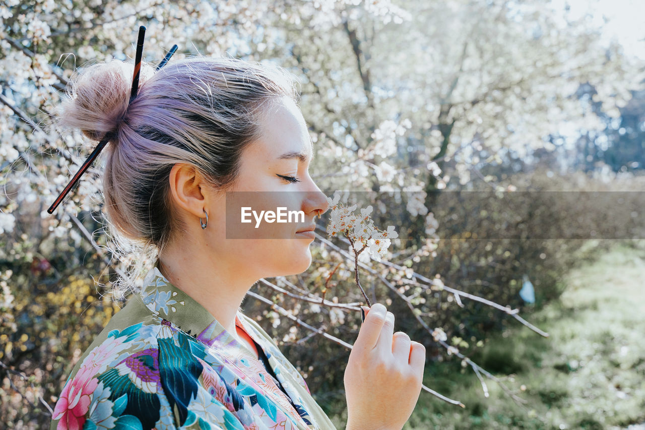 Side view of young female in traditional kimono holding blossoming flower sprig in garden on sunny day looking at camera