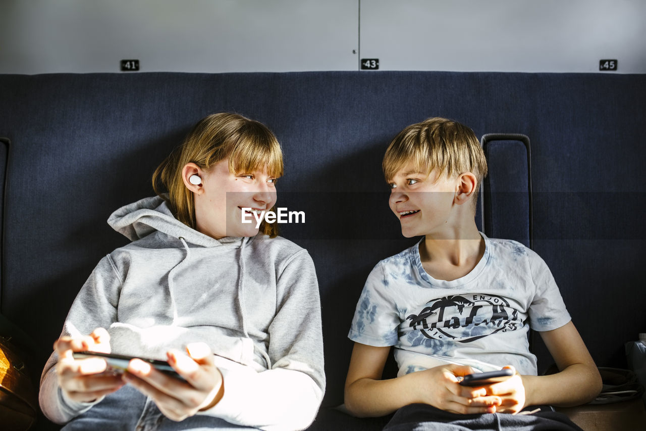 Smiling brother and sister with smart phones looking at each other while traveling in train