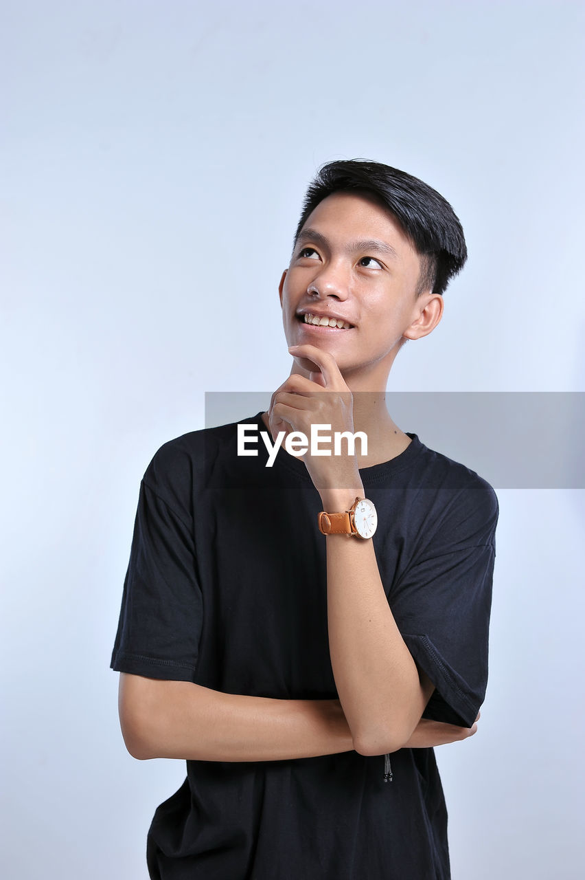 Handsome young asian boy wearing black t-shirt and wristwatch with hand on chin thinking about question, pensive expression. smiling with thoughtful face. doubt concept