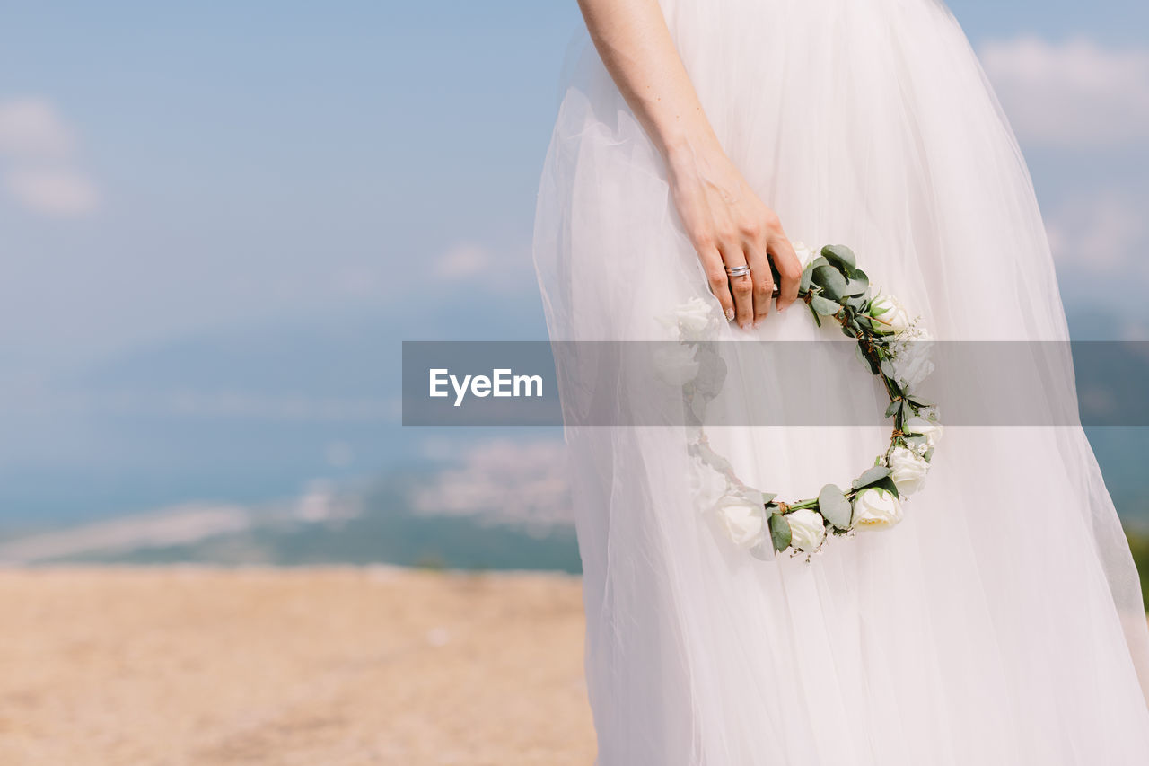 Midsection of bride holding wreath against sky