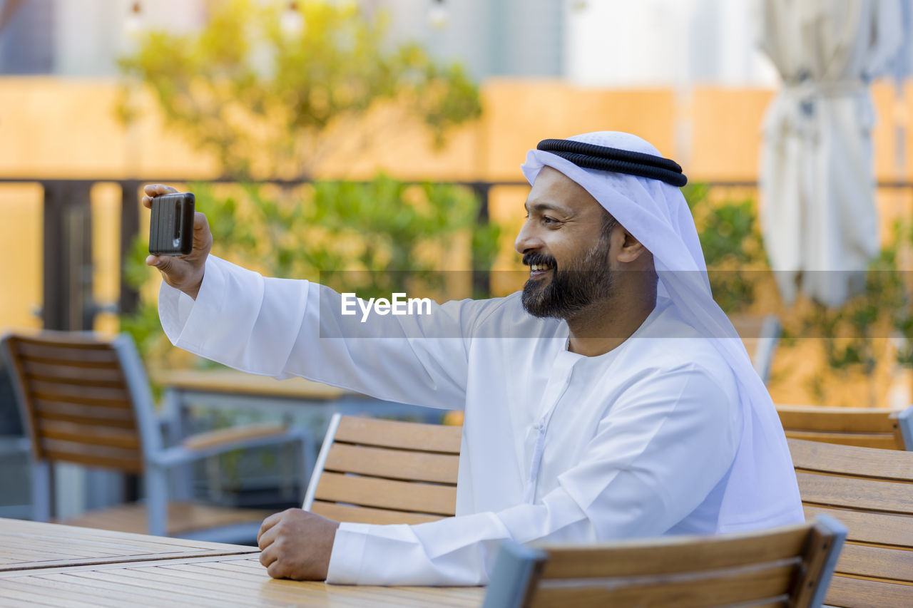 Man wearing traditional clothing taking selfie at outdoor cafe