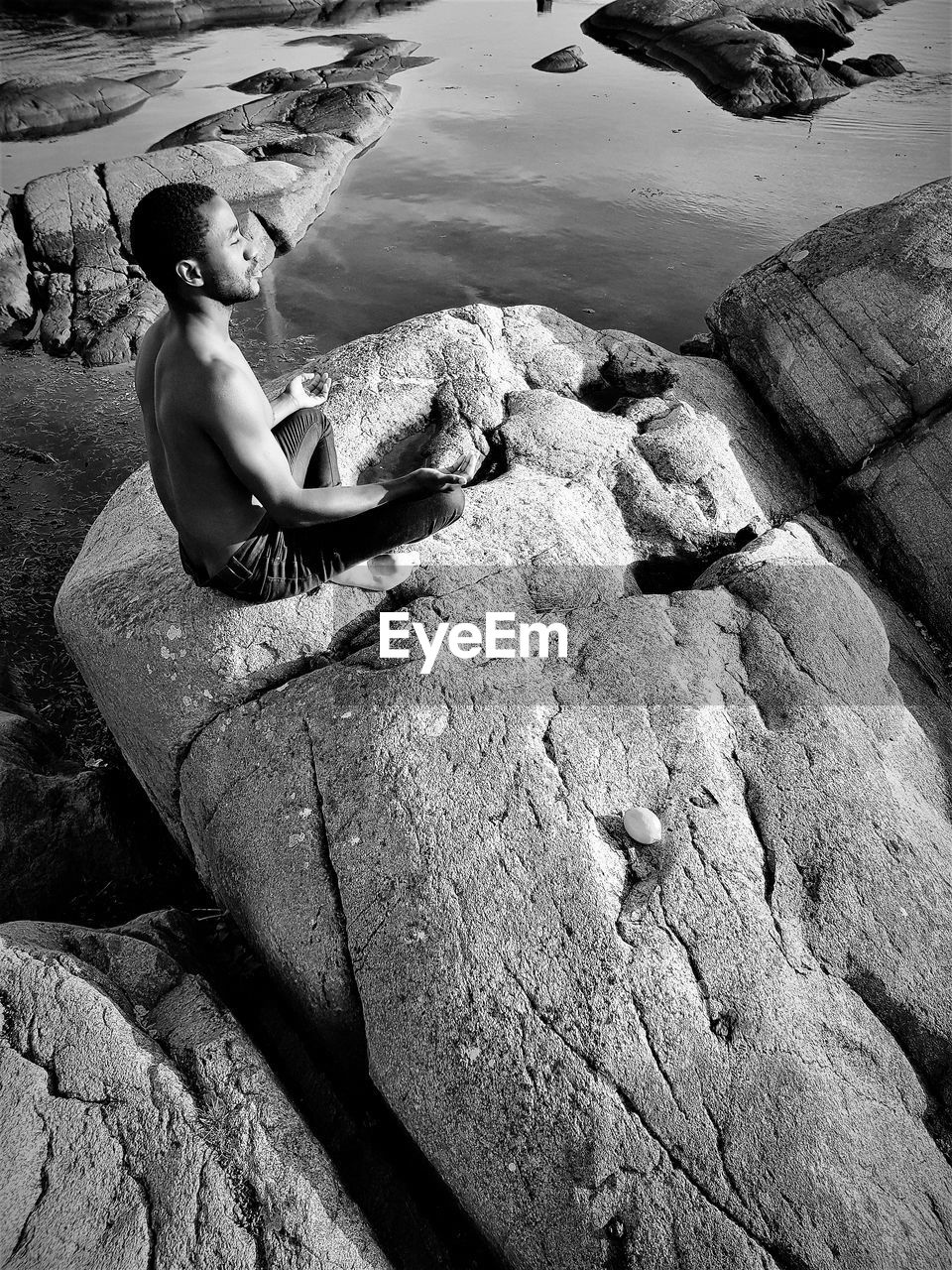 High angle view of shirtless young man mediating while sitting on rock at beach