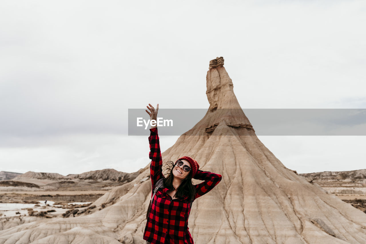 Spain, navarre, portrait of female tourist standing with raised arms in front of sandstone rock formation in bardenas reales