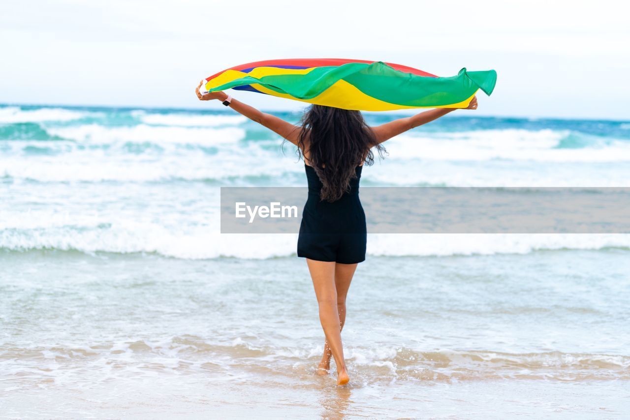 water, sea, beach, land, one person, wave, adult, umbrella, nature, horizon over water, women, sports, vacation, motion, trip, sky, clothing, surfing equipment, protection, leisure activity, surfing, holiday, wet, ocean, water sports, horizon, summer, surfboard, lifestyles, beauty in nature, young adult, shore, standing, full length, day, relaxation, wind, sand, holding, swimwear, outdoors, rear view, enjoyment, coast, female, environment, walking, travel destinations, happiness, emotion, human leg, security, carefree, rain, idyllic, person, parasol, travel, scenics - nature, vitality, water's edge, multi colored, solitude